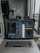 A laptop displaying code on the screen with various open applications is placed on a table in a modern workspace. The background features a concrete wall and windows offering a view of a neighboring building and street below.