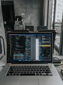 A laptop displaying code on the screen with various open applications is placed on a table in a modern workspace. The background features a concrete wall and windows offering a view of a neighboring building and street below.