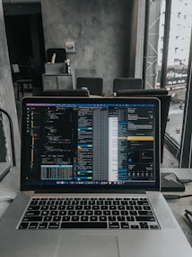 A laptop displaying code on the screen with various open applications is placed on a table in a modern workspace. The background features a concrete wall and windows offering a view of a neighboring building and street below.