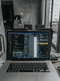 A laptop displaying code on the screen with various open applications is placed on a table in a modern workspace. The background features a concrete wall and windows offering a view of a neighboring building and street below.
