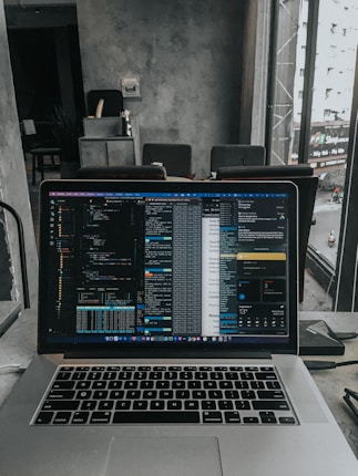 A laptop displaying code on the screen with various open applications is placed on a table in a modern workspace. The background features a concrete wall and windows offering a view of a neighboring building and street below.