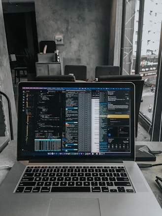 A laptop displaying code on the screen with various open applications is placed on a table in a modern workspace. The background features a concrete wall and windows offering a view of a neighboring building and street below.