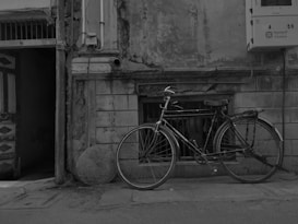 A vintage bicycle is positioned against an old, weathered building with peeling paint and visible brickwork. The street is narrow, with a slightly open wooden door leading to a dimly lit interior. An electrical box with wires is mounted on the wall above the bicycle.