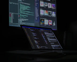 A dimly lit room features a desktop monitor and a laptop on a desk, both displaying lines of code and digital graphics. The primary focus is on programming and software development, with a dark, tech-centric theme.