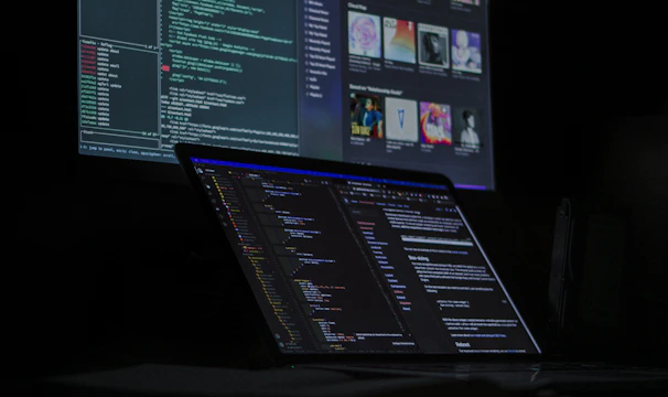 A dimly lit room features a desktop monitor and a laptop on a desk, both displaying lines of code and digital graphics. The primary focus is on programming and software development, with a dark, tech-centric theme.