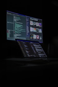 A dimly lit room features a desktop monitor and a laptop on a desk, both displaying lines of code and digital graphics. The primary focus is on programming and software development, with a dark, tech-centric theme.