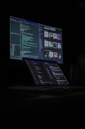 A dimly lit room features a desktop monitor and a laptop on a desk, both displaying lines of code and digital graphics. The primary focus is on programming and software development, with a dark, tech-centric theme.