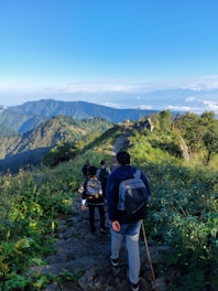 A group of travelers hiking through a lush green mountain trail under a bright blue sky.