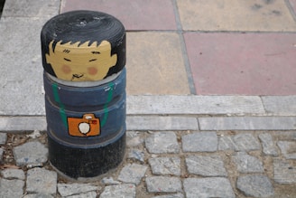 A painted bollard on a cobblestone street resembles a person with a cartoon face and black hair. The bollard is also painted with an image of an orange camera hung around the 'neck'. The surrounding area includes stone paving and tiles in shades of brown and red.