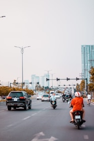 A busy urban street with several motorcycles and cars traveling in one direction. A person is riding a motorbike in an orange shirt and white helmet in the foreground. Tall buildings and traffic lights are visible in the distance, and the sky is overcast.