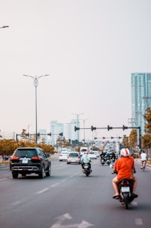 A busy urban street with several motorcycles and cars traveling in one direction. A person is riding a motorbike in an orange shirt and white helmet in the foreground. Tall buildings and traffic lights are visible in the distance, and the sky is overcast.