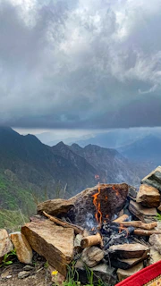 A campfire glowing orange under a stormy sky, surrounded by survival gear and maps.