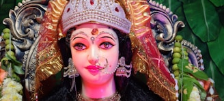 A detailed and vibrant depiction of a goddess figure adorned with an elaborately decorated crown and intricate jewelry. The face is painted with expressive eyes and a serene expression, surrounded by colorful fabrics and ornate decorations. In the background, green leaves and floral elements add to the richness and cultural ambiance.