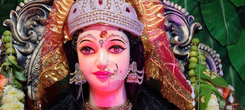 A detailed and vibrant depiction of a goddess figure adorned with an elaborately decorated crown and intricate jewelry. The face is painted with expressive eyes and a serene expression, surrounded by colorful fabrics and ornate decorations. In the background, green leaves and floral elements add to the richness and cultural ambiance.