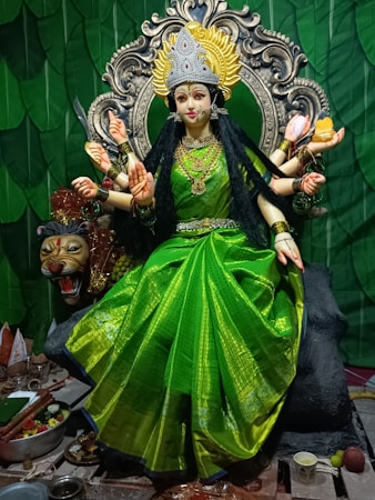 A detailed statue of a goddess with multiple arms, adorned with intricate jewelry and a bright green saree. The goddess has a serene expression and sits on a throne with an ornate backdrop featuring a large decorative halo. A fierce-looking lion figure is positioned beside her, and various items are placed at the base, including offerings and ritual objects.