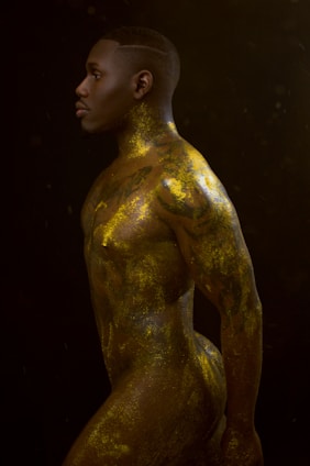 A side profile of a person with a muscular build covered in gold body paint. The image features dramatic lighting that highlights the contours of the body against a dark background, creating a striking visual contrast.