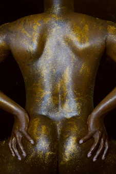 A person is posed with their back facing the viewer, covered completely in glittering gold body paint. The person's hands are resting on their lower back, highlighting the contours and muscles with a shimmering effect.