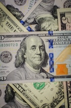 A close-up view of a pile of various denominations of US dollar bills, centered around a prominent hundred-dollar note featuring Benjamin Franklin. The banknotes display different colors and intricate details, with visible seals and serial numbers.