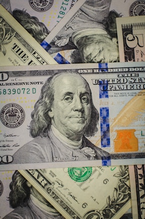 A close-up view of a pile of various denominations of US dollar bills, centered around a prominent hundred-dollar note featuring Benjamin Franklin. The banknotes display different colors and intricate details, with visible seals and serial numbers.