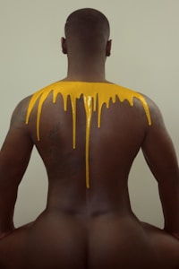 A person with their back facing the camera, with a vibrant yellow liquid appearing to drip down from their shoulders. The background is a neutral light color, highlighting the contrast between the person's skin tone and the yellow substance.