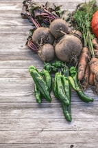 A rustic arrangement of freshly harvested vegetables including beets, green peppers, carrots, and a hint of a pumpkin. The produce is placed on a weathered wooden surface, with dirt still clinging to some of the roots and leaves, suggesting a recent harvest.