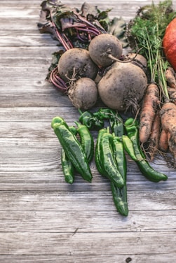 A rustic arrangement of freshly harvested vegetables including beets, green peppers, carrots, and a hint of a pumpkin. The produce is placed on a weathered wooden surface, with dirt still clinging to some of the roots and leaves, suggesting a recent harvest.