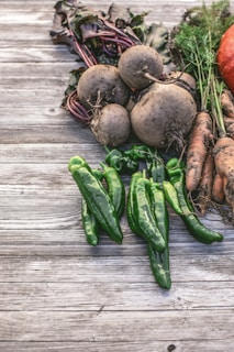 A rustic arrangement of freshly harvested vegetables including beets, green peppers, carrots, and a hint of a pumpkin. The produce is placed on a weathered wooden surface, with dirt still clinging to some of the roots and leaves, suggesting a recent harvest.