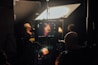 A film or photography studio setup with a focus on a monitor displaying a close-up of a person's face. Multiple individuals are present, engaged in the production process. Studio lighting and equipment are visible, creating an atmosphere of professional film or photo shoot environment.