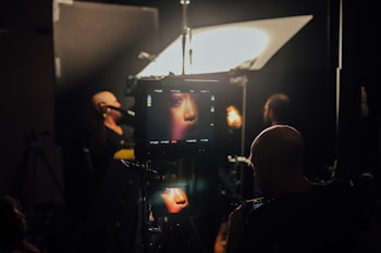 A film or photography studio setup with a focus on a monitor displaying a close-up of a person's face. Multiple individuals are present, engaged in the production process. Studio lighting and equipment are visible, creating an atmosphere of professional film or photo shoot environment.