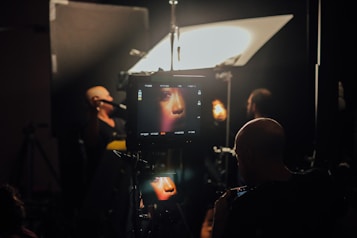 A film or photography studio setup with a focus on a monitor displaying a close-up of a person's face. Multiple individuals are present, engaged in the production process. Studio lighting and equipment are visible, creating an atmosphere of professional film or photo shoot environment.