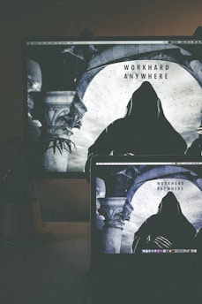 A dimly lit workspace features a laptop in the foreground, displaying an image of a hooded figure standing beneath an arched structure, with the words 'WORK HARD ANYWHERE' above. In the background, a larger monitor shows the same image, creating a mirrored effect. The setting includes elements such as a plant and possibly a mug or container on the side.