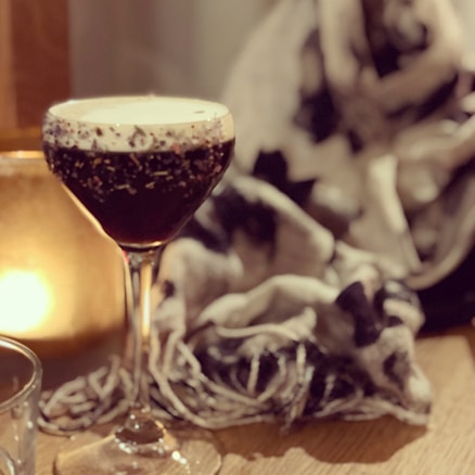 A dark cocktail in a delicate glass with a frothy cream layer on top, set beside a soft, blurred background with warm lighting. The scene includes a textured scarf and a soft glow from a light source, creating a cozy and intimate atmosphere.