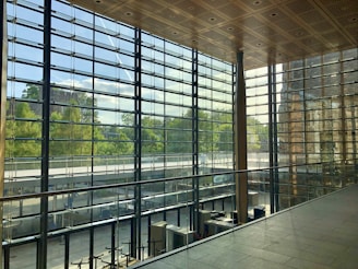 A spacious interior with large floor-to-ceiling glass windows offering a view of lush greenery outside. The ceiling features a wooden grid pattern, and the area is elegantly designed with metal and glass railing. The natural light creates a bright and airy atmosphere within the space.