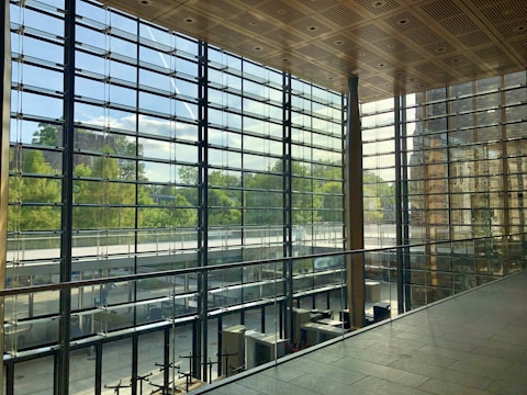 A spacious interior with large floor-to-ceiling glass windows offering a view of lush greenery outside. The ceiling features a wooden grid pattern, and the area is elegantly designed with metal and glass railing. The natural light creates a bright and airy atmosphere within the space.