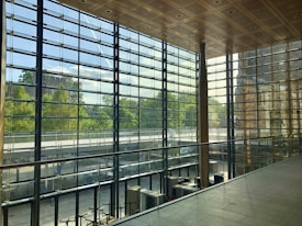 A spacious interior with large floor-to-ceiling glass windows offering a view of lush greenery outside. The ceiling features a wooden grid pattern, and the area is elegantly designed with metal and glass railing. The natural light creates a bright and airy atmosphere within the space.