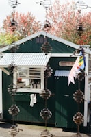 A small green building with a white corrugated roof stands outdoors surrounded by autumn trees with red foliage. The building has a window with several items on the sill, and a white towel and spray bottle are hanging on the wall. The word 'Nursery' is displayed on a sign near the entrance, and colorful flags are attached to the side. The foreground features a vertical chain-like structure, adding depth to the image.