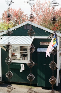 A small green building with a white corrugated roof stands outdoors surrounded by autumn trees with red foliage. The building has a window with several items on the sill, and a white towel and spray bottle are hanging on the wall. The word 'Nursery' is displayed on a sign near the entrance, and colorful flags are attached to the side. The foreground features a vertical chain-like structure, adding depth to the image.