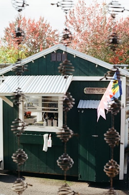 A small green building with a white corrugated roof stands outdoors surrounded by autumn trees with red foliage. The building has a window with several items on the sill, and a white towel and spray bottle are hanging on the wall. The word 'Nursery' is displayed on a sign near the entrance, and colorful flags are attached to the side. The foreground features a vertical chain-like structure, adding depth to the image.