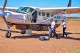 A small aircraft is parked on a dirt runway with two pilots standing nearby. One pilot is carrying luggage and the other is speaking on a phone. The aircraft door is open, and the plane displays the registration 5Y-SLB.