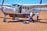 A small aircraft is parked on a dirt runway with two pilots standing nearby. One pilot is carrying luggage and the other is speaking on a phone. The aircraft door is open, and the plane displays the registration 5Y-SLB.