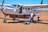 A small aircraft is parked on a dirt runway with two pilots standing nearby. One pilot is carrying luggage and the other is speaking on a phone. The aircraft door is open, and the plane displays the registration 5Y-SLB.