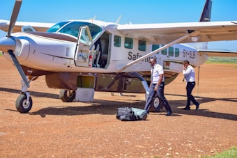 A small aircraft is parked on a dirt runway with two pilots standing nearby. One pilot is carrying luggage and the other is speaking on a phone. The aircraft door is open, and the plane displays the registration 5Y-SLB.