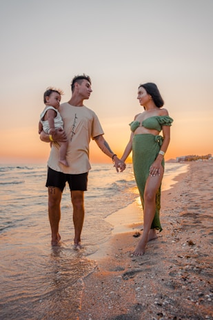 A family stands on a sandy beach with gentle waves lapping at their feet. The father holds a small child while the mother holds the father's hand. The sunset creates a warm, golden glow on the horizon.