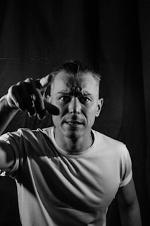 A black and white portrait features a man wearing a plain t-shirt, with a focused and intense expression. He points directly at the camera, creating a sense of direct engagement. The background is plain and dark, adding to the dramatic effect of the lighting and shadows.