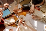 A group of people is gathered around a wooden table filled with open books, papers, and stationery. The table also has various items like a radio, calculator, and a whole orange. One person is writing while others point at the papers, indicating a collaborative study or brainstorming session. The sunlight casts shadows, adding a warm ambiance.