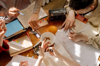 A group of people is gathered around a wooden table filled with open books, papers, and stationery. The table also has various items like a radio, calculator, and a whole orange. One person is writing while others point at the papers, indicating a collaborative study or brainstorming session. The sunlight casts shadows, adding a warm ambiance.