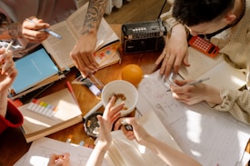 A group of people is gathered around a wooden table filled with open books, papers, and stationery. The table also has various items like a radio, calculator, and a whole orange. One person is writing while others point at the papers, indicating a collaborative study or brainstorming session. The sunlight casts shadows, adding a warm ambiance.