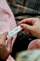 A person is holding a home pregnancy test with two visible lines, indicating a positive result. The background shows a soft and warm environment with a pink textured blanket and a plaid pattern fabric.