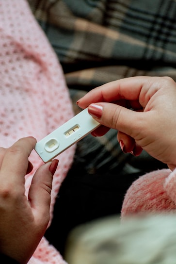 A person is holding a home pregnancy test with two visible lines, indicating a positive result. The background shows a soft and warm environment with a pink textured blanket and a plaid pattern fabric.