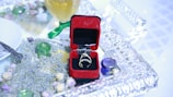 A red velvet jewelry box containing a ring rests on an ornate silver tray. Surrounding the box are dried lavender, small white roses, and decorative glass items. A golden beverage sits nearby in a goblet, next to white tableware and a silver utensil.